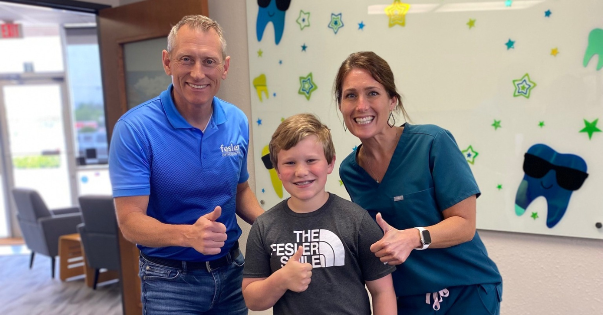 A young boy stands between a man and woman in Fesler Orthodontic office, all three looking at the camera with thumbs up and friendly expressions.